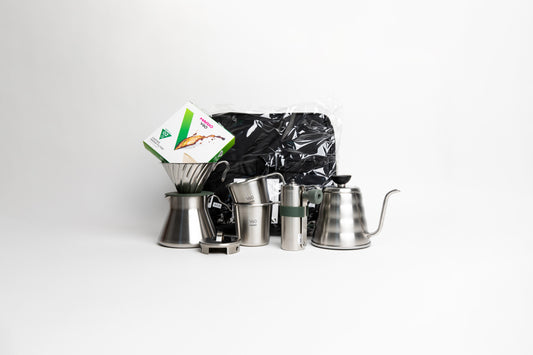 stainless steel kettle, metal cone coffee dripper with a olive green base on top of a stainless steel coffee sever pot, stainless steel lid, with metal coffee hand grinder and two metal cups mugs, box of filters and a black bag. on a white background