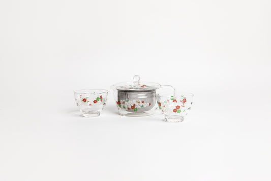 All glass tapered tea pot with flat bottom, all glass lid and handle, and metal mesh filter basket insert. decorated glass with a minimal flower design  with match small all glass tea cups with flower design. set on a white background.