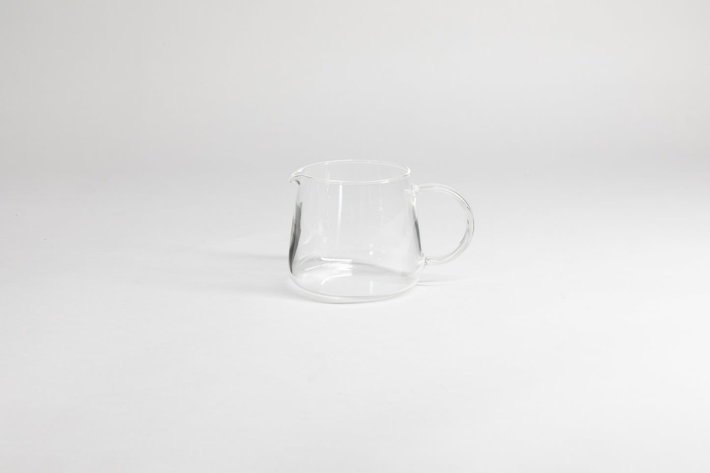 Round glass server with glass handle and fluted spout. On a light grey background.