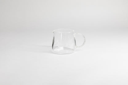 Round glass server with glass handle and fluted spout. On a light grey background.