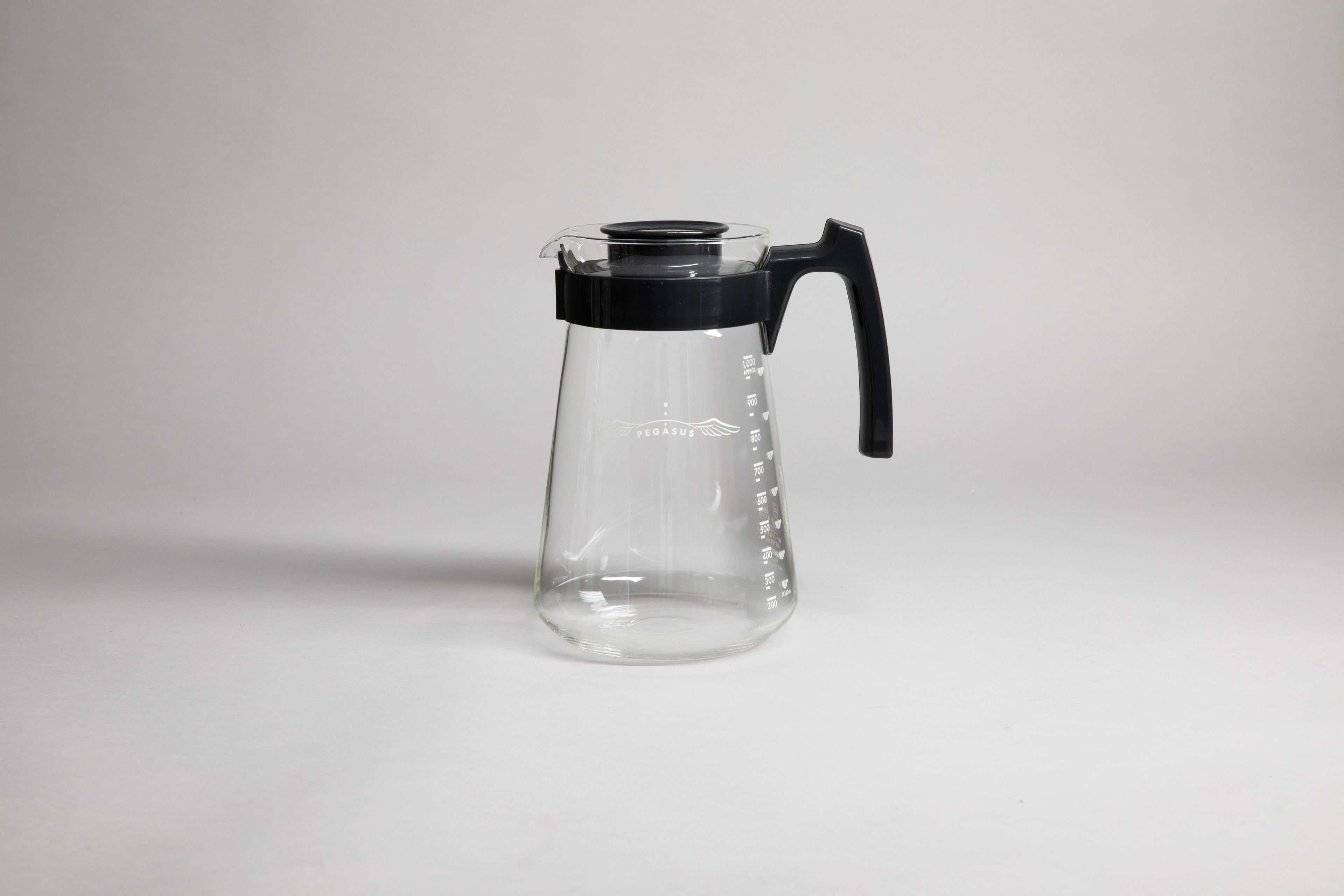 Glass coffee server with white text and level markings with black hard plastic handle and lid on a light grey background.