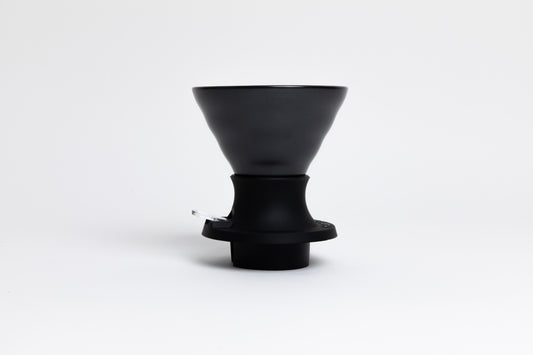 Cone-shaped matte black ceramic dripper seated in a black silicone base with clear plastic push switch on a white background.