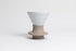 Cone-shaped matte white ceramic dripper seated in a light tan silicone base with clear plastic push switch on a white background.