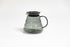 All glass transparent gray beehive shaped coffee server with glass handle and glass lid with dark plastic rubber gasket on white backdrop.