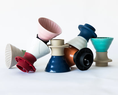 A colorful assortment of V60 ceramic cone coffee drippers with various silicone  bases, arranged in a pile on a white background. 
