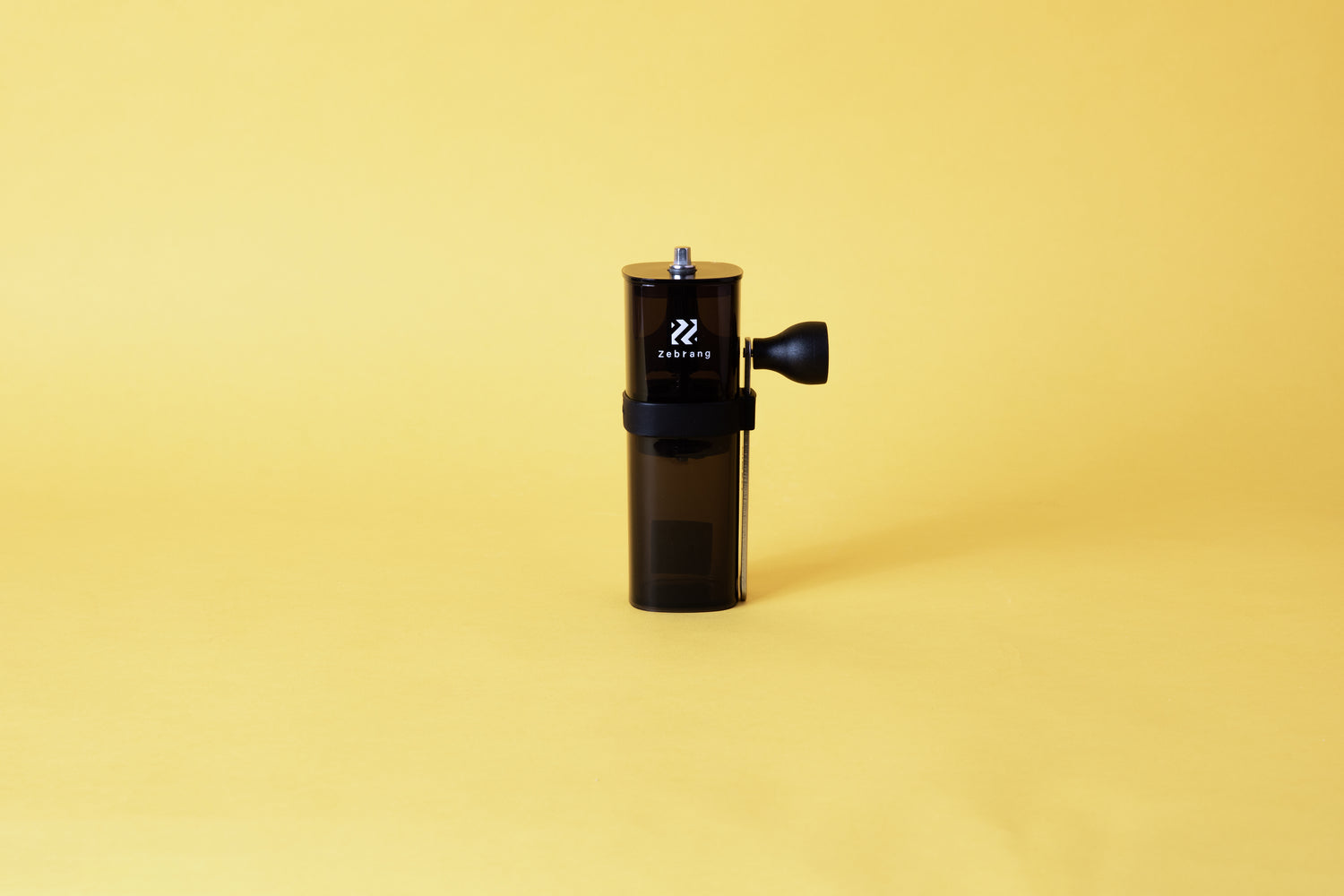 a transparent black cylindrical coffee mill with a middle band for storing the detachable handle.  Set on yellow background.