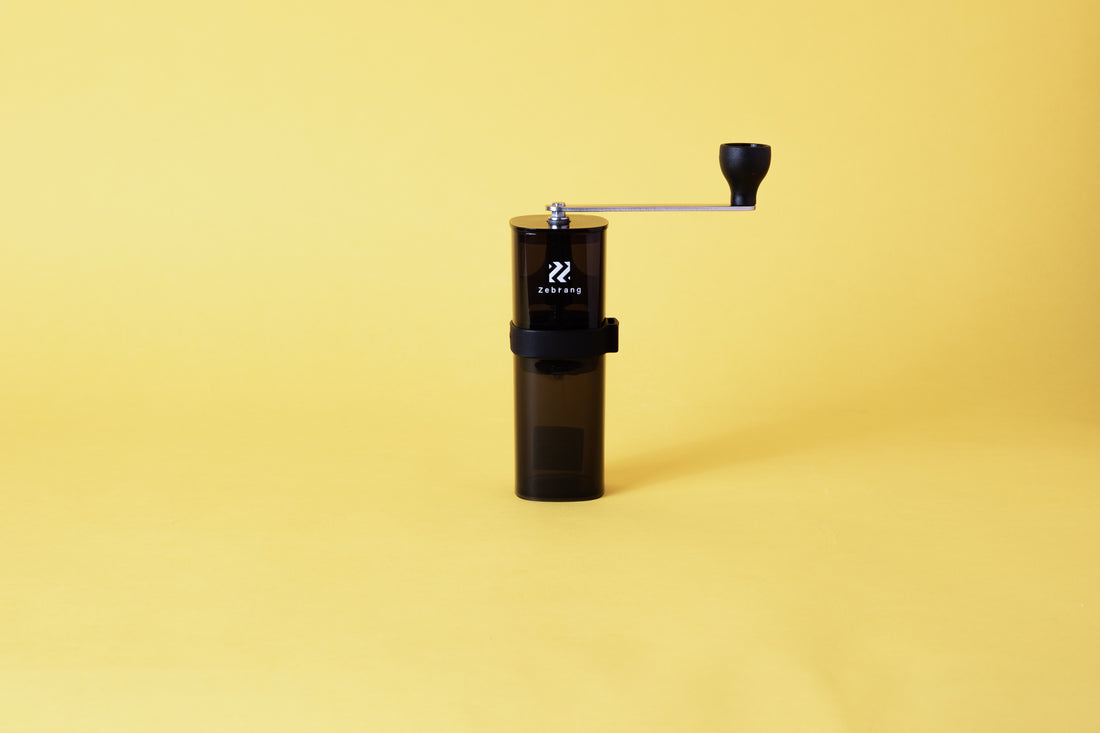 a transparent black cylindrical coffee mill with a metal handle bar and black knob.  Set on yellow background.