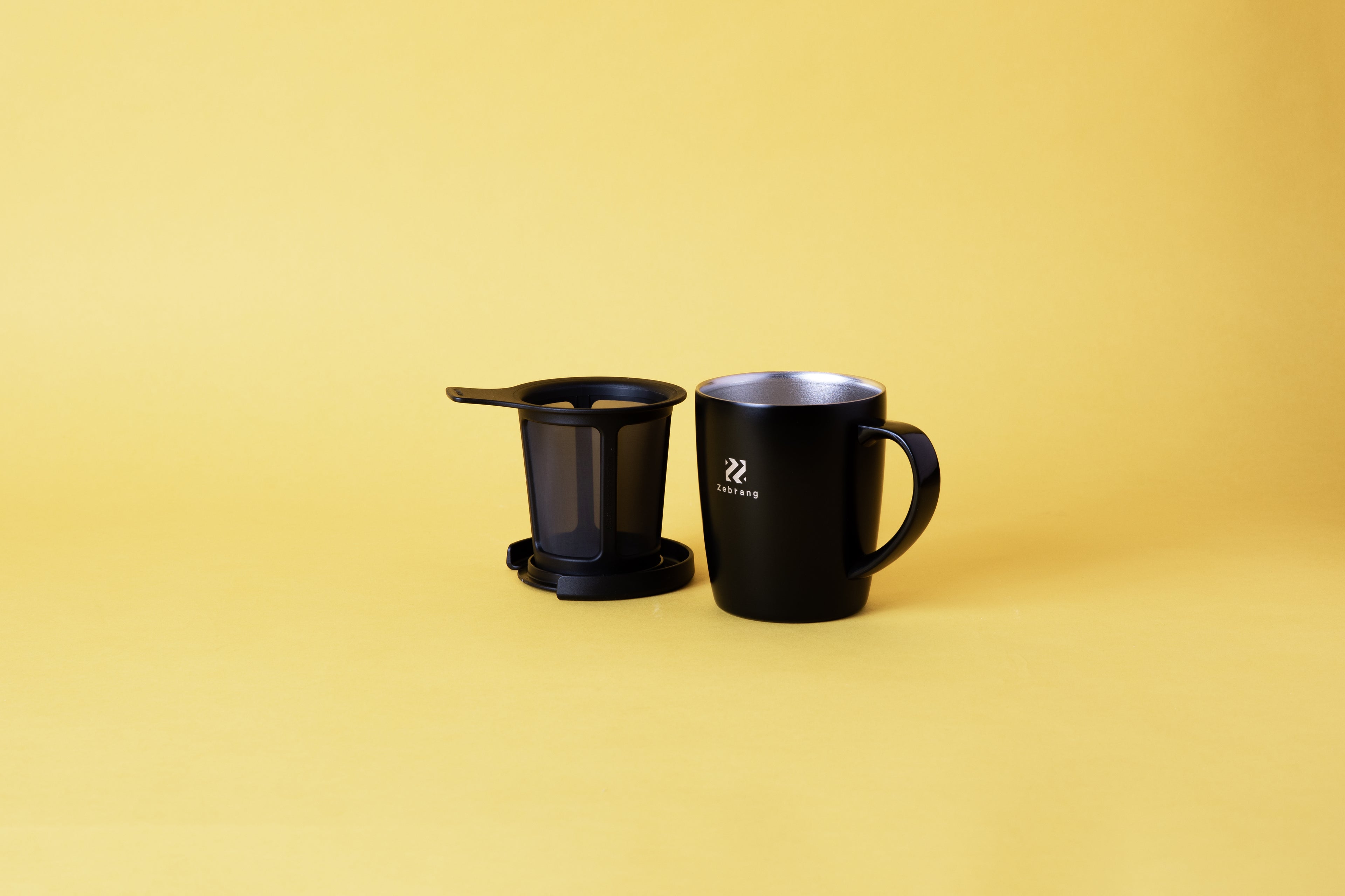 Black mug with handle, exposing inside of the mug which is stainless steel. Coffee filter basket that can be placed inside the mug. The lid of the mug is used as a coffee basket drip tray. set on yellow background