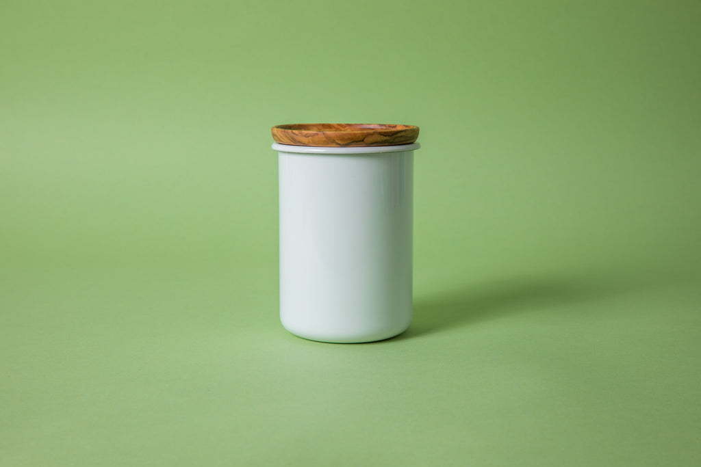 Cylindrical white enamel container with olive wood lid and green background.