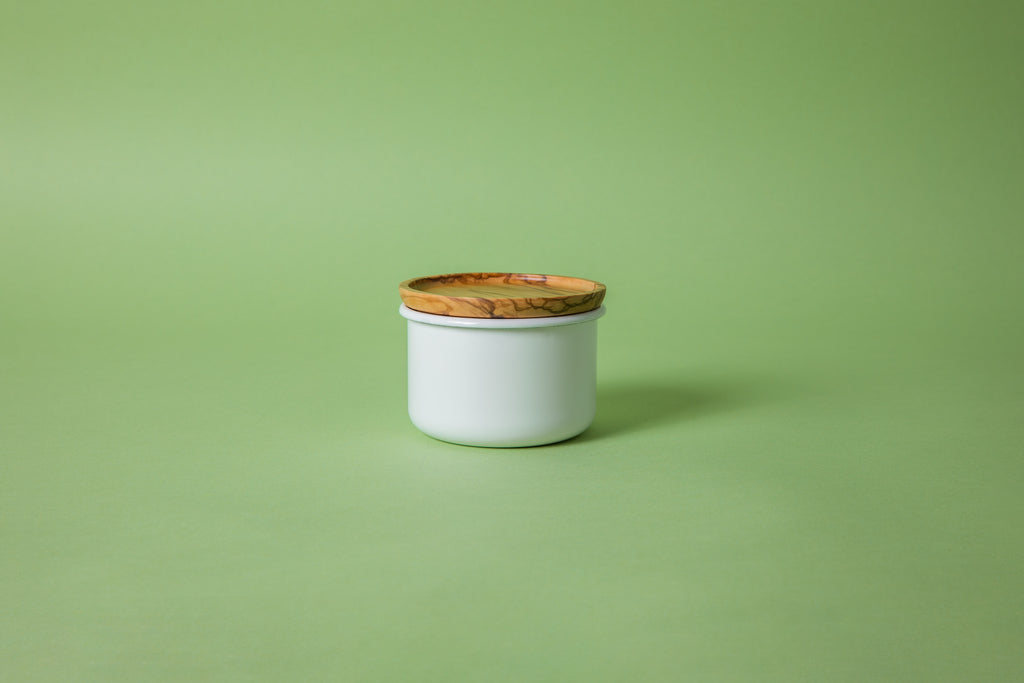 Cylindrical white enamel container with olive wood lid and green background.