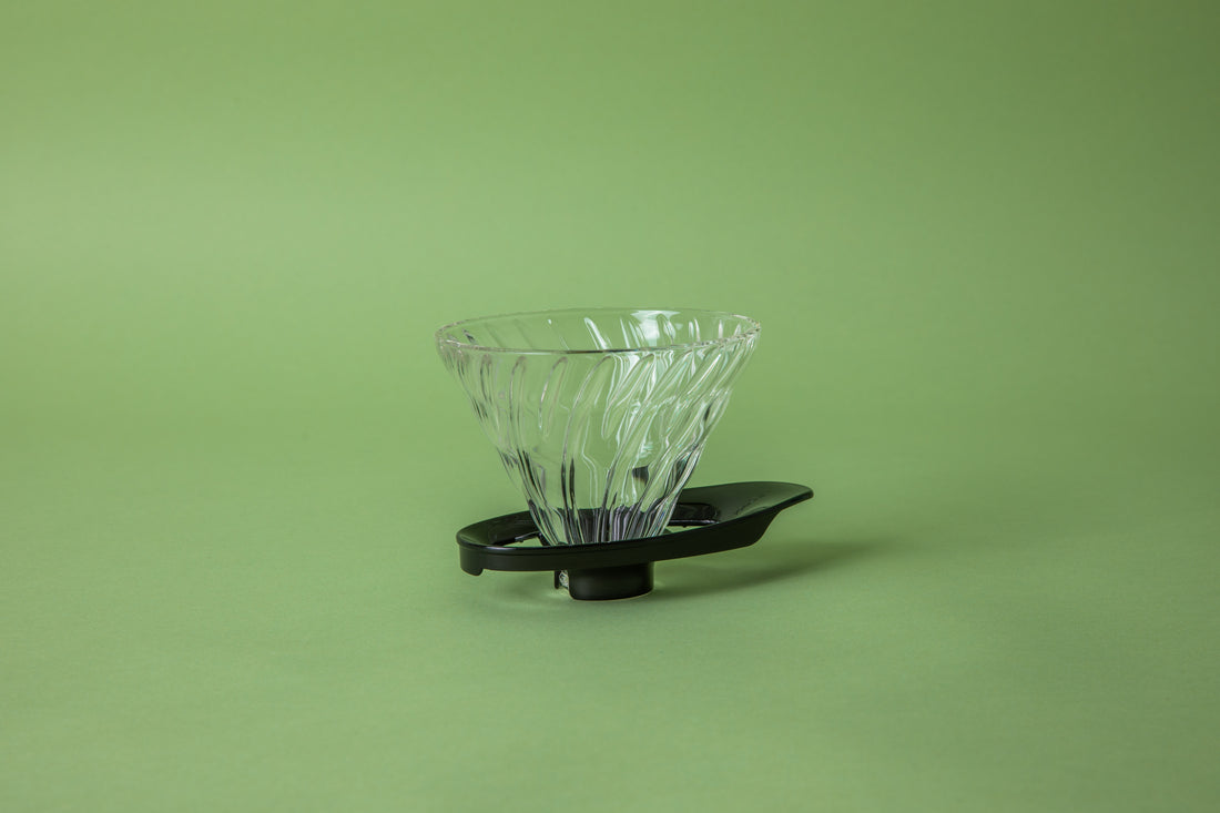 Clear transparent cone shaped coffee dripper with ribbed spiral. on a black plastic hollow base. Base shaped like a surfboard. Set on green background. 