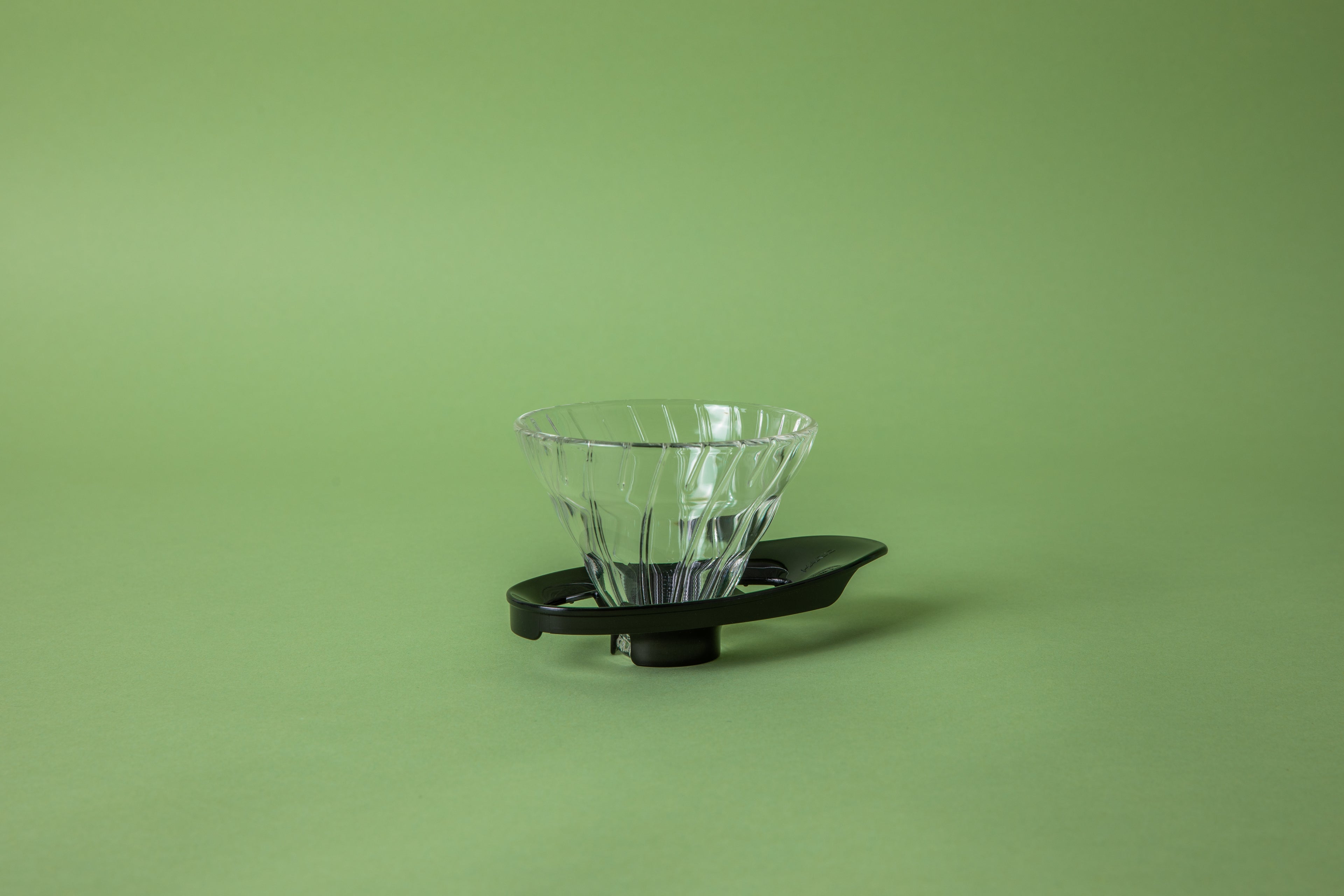 Clear transparent cone shaped coffee dripper with ribbed spiral. on a black plastic hollow base. Base shaped like a surfboard. Set on green background. 