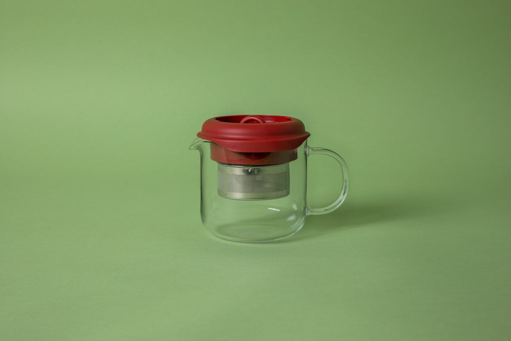 glass beaker with glass handle with red silicone lid, the lid knob is pushed down exposing staineless steel filter basket. set on green background. 