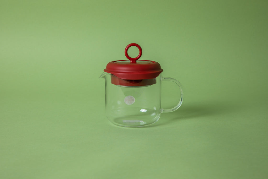 Glass beaker with glass handle with red silicone lid and a circular red knob. set on green background
