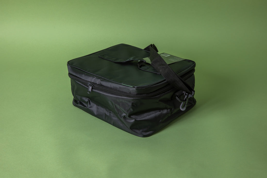 Square black bag with black strap hooked on black plastic clip. Set on green background.