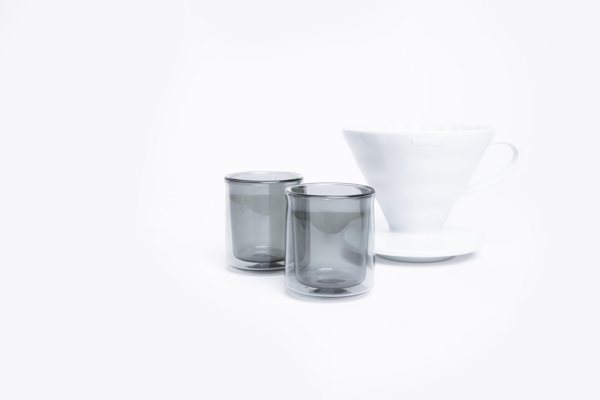 Two smoked glass tumblers and a white cone dripper with handle on a white background