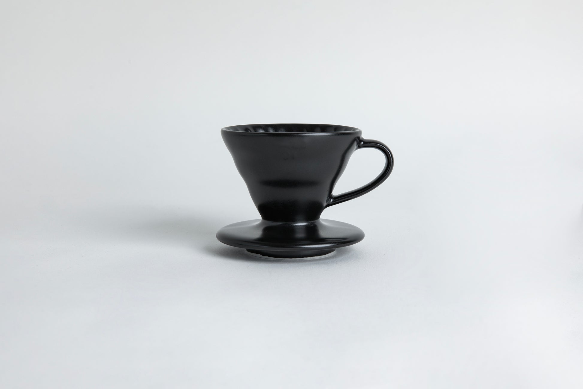 matte black cone shaped ceramic coffee dripper with handle and round base. Spiral ribbed on the inside cone. Set on light gray background