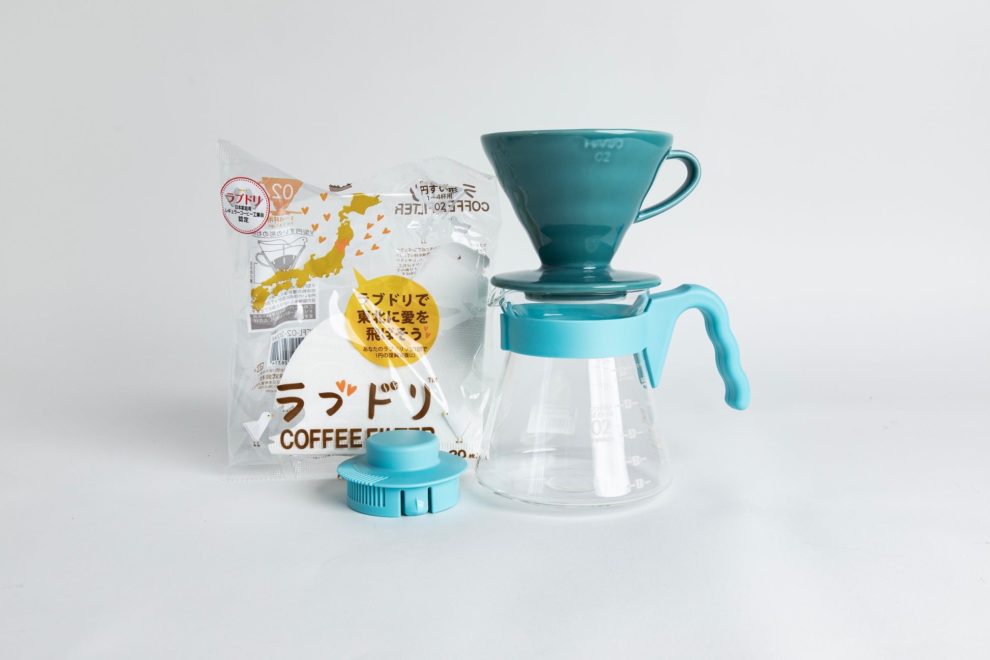Teal HARIO 02 cone dripper on a glass carafe with a light teal handle, shown with paper filters and a matching teal lid on a light gray background.