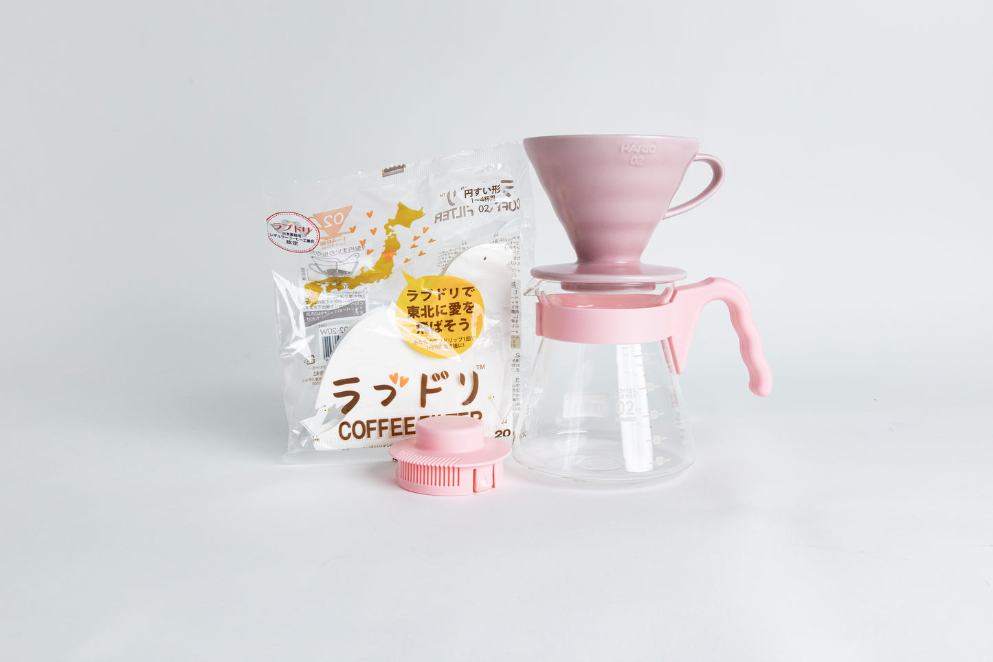 Light pink HARIO 02 cone dripper on a glass carafe with a pink handle, shown with paper filters and a matching pink lid on a light gray background.