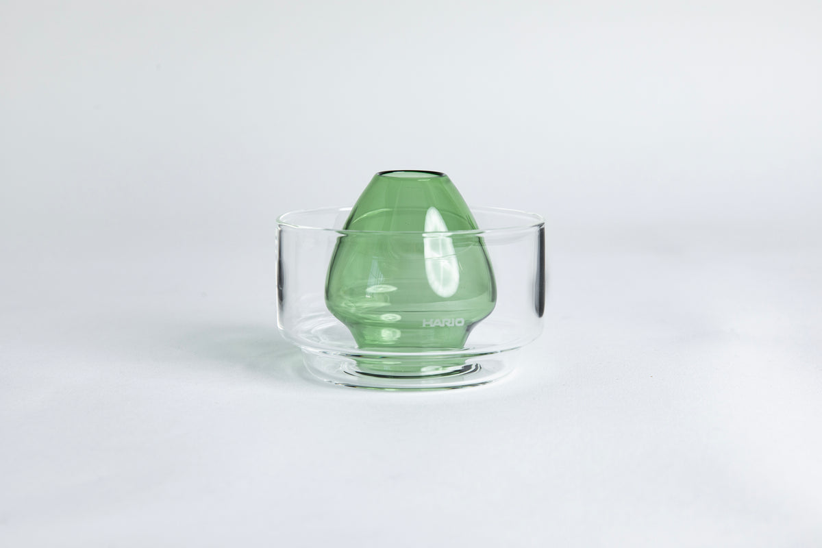 Dome-shaped green clear glass Matcha whisk holder with a smooth, tapered form, placed inside a clear glass base. Displayed on a white background with 'Hario' branding
