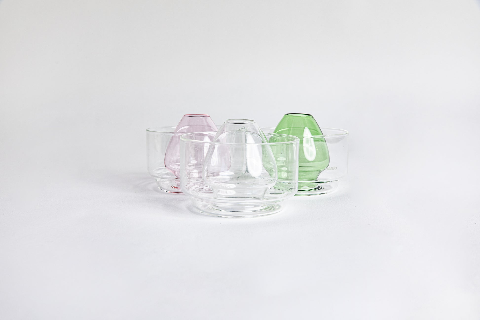 Three side by side images of a dome-shaped in the colors clear, pink and green clear glass matcha whisk holder with a smooth, tapered form, placed inside a clear glass base. Displayed on a white background with 'Hario' branding