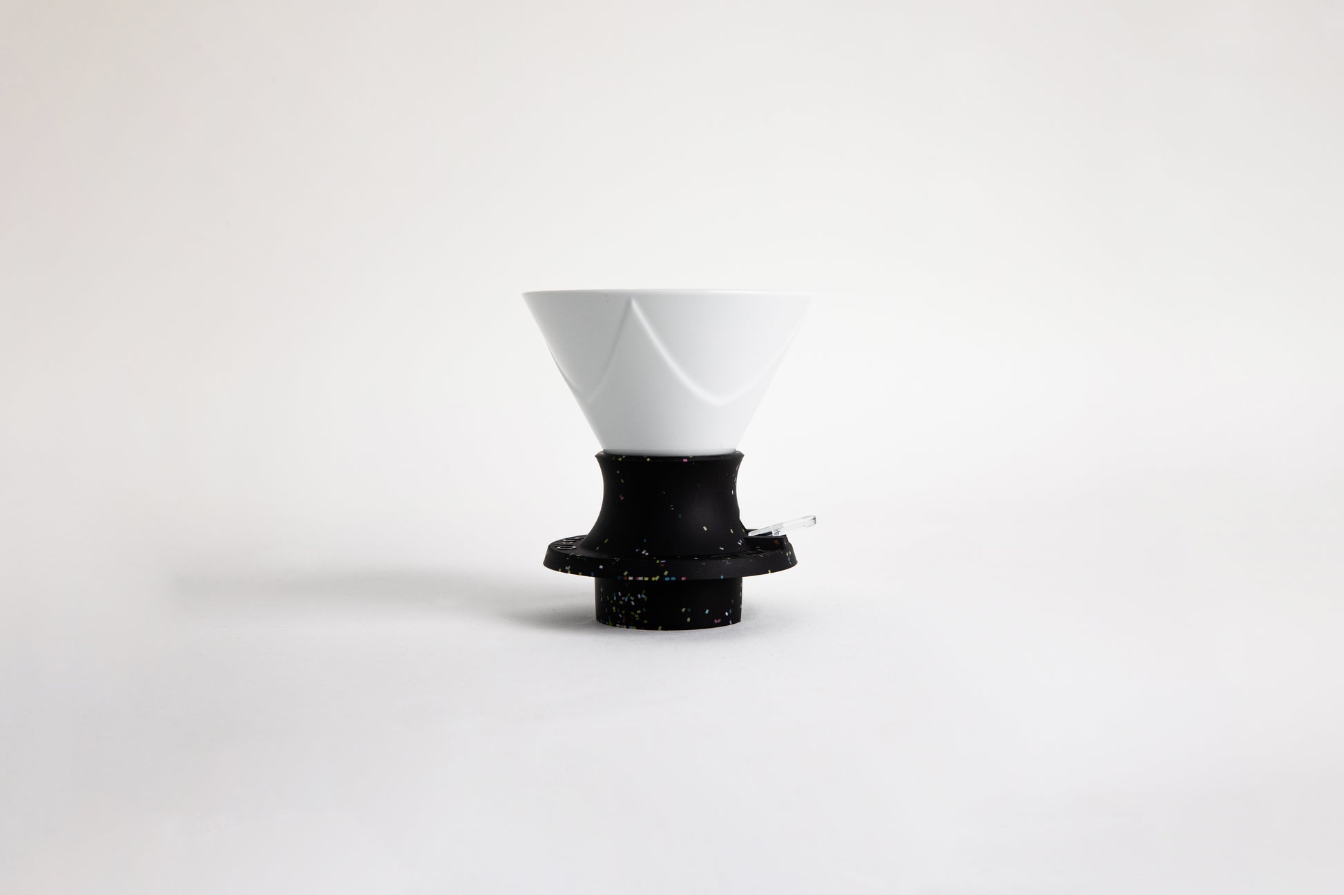 White ceramic cone-shaped dripper with flowing Bezier-style ridges, set in a black silicone base that features rainbow speckles. A clear plastic switch. set on a white background.