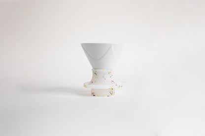 White ceramic cone-shaped dripper with flowing Bezier-style ridges, set in a white silicone base that features rainbow speckles. A clear plastic switch. set on a white background.