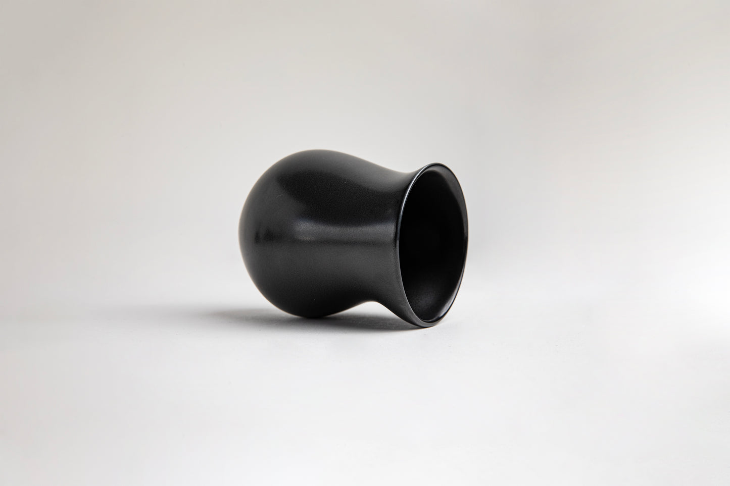 side view matte black ceramic vessel with a curvy, tulip-like silhouette. The surface has a smooth texture, on a light gray background.