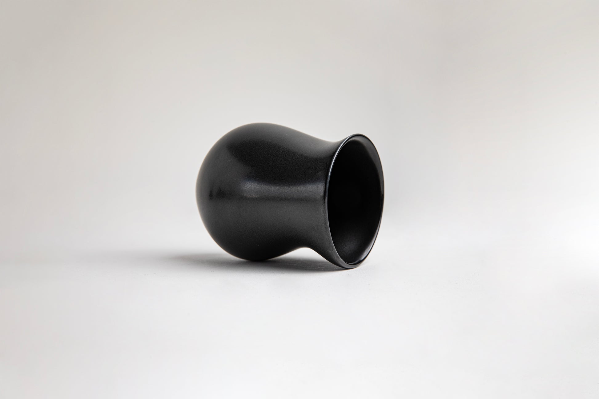 side view matte black ceramic vessel with a curvy, tulip-like silhouette. The surface has a smooth texture, on a light gray background.
