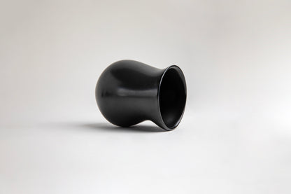 side view matte black ceramic vessel with a curvy, tulip-like silhouette. The surface has a smooth texture, on a light gray background.
