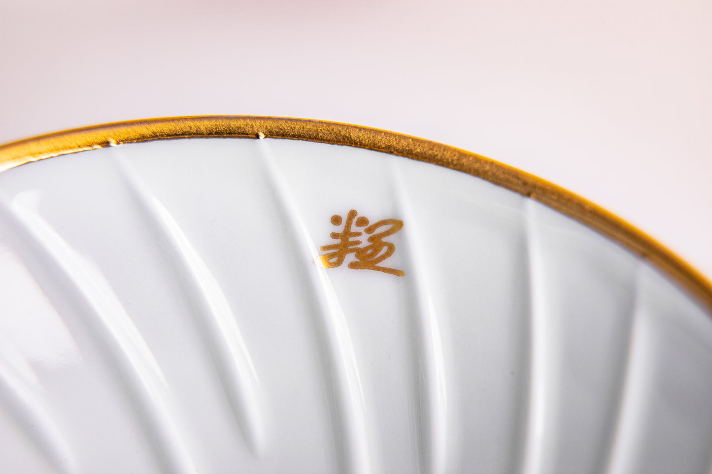 Close-up of a white ceramic dripper with gold rim and gold text on a light pink background