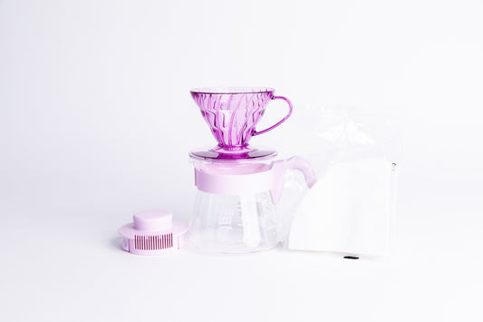 Transparent Purple plastic coffee cone dripper on glass server with light purple handle and lid with a pack of white filters on a white background
