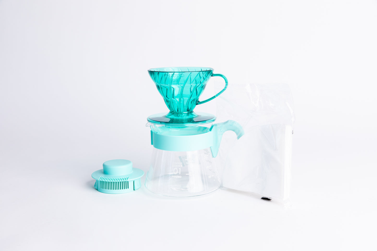 Transparent teal plastic coffee cone dripper on glass server with light teal handle and lid with a pack of white filters on a white background