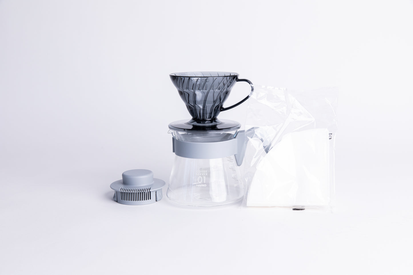 Transparent gray plastic coffee cone dripper on glass server with light gray handle and lid with a pack of white filters on a white background