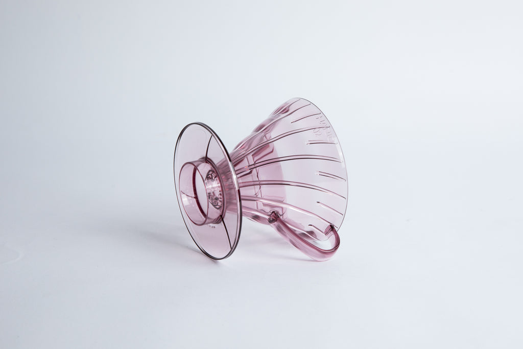 set on it's side the Size 02 Plastic V60 Dripper in the color transparent blushy mauve (Stardust) with a cylindrical base and curved handle. Set on a white background