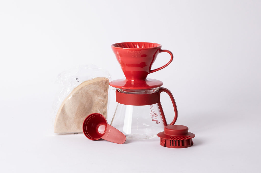 Cellophane wrapped Brown Coffee filter, plastic red coffee scoop, 360ml glass pitcher with a red collar and handle, 01 size red ceramic V60 Dripper, and a red cap for the pitcher. set on a white background