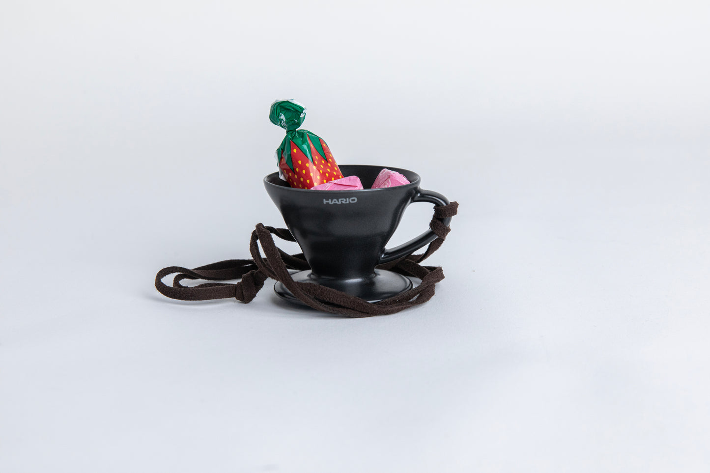 matte black cone shaped ceramic coffee dripper  cup with handle and round base.  with a brown rope tied on the handle. with  small pieces of candy inside showcasing the size of the small cup Set on white background