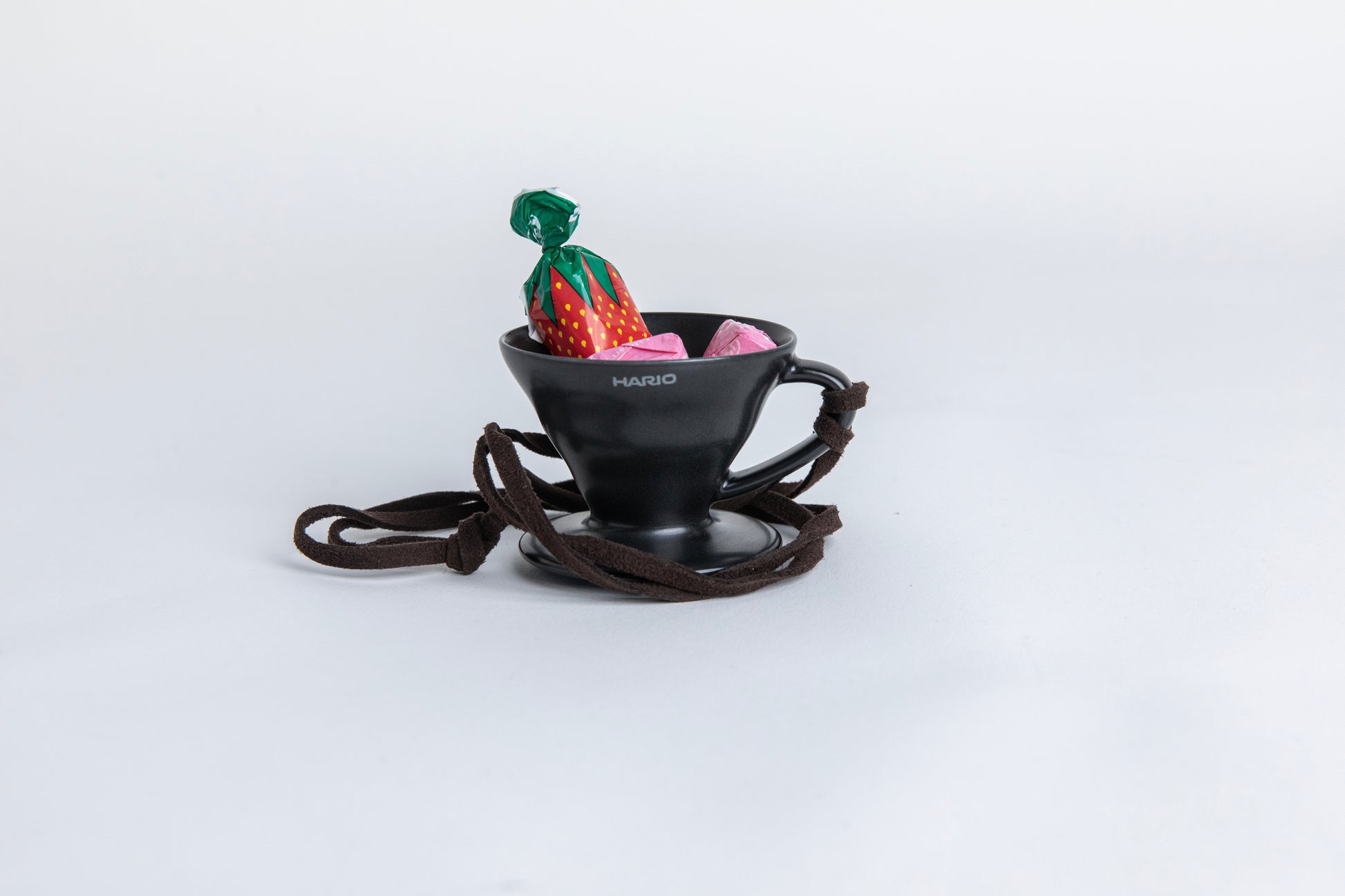 matte black cone shaped ceramic coffee dripper  cup with handle and round base.  with a brown rope tied on the handle. with  small pieces of candy inside showcasing the size of the small cup Set on white background