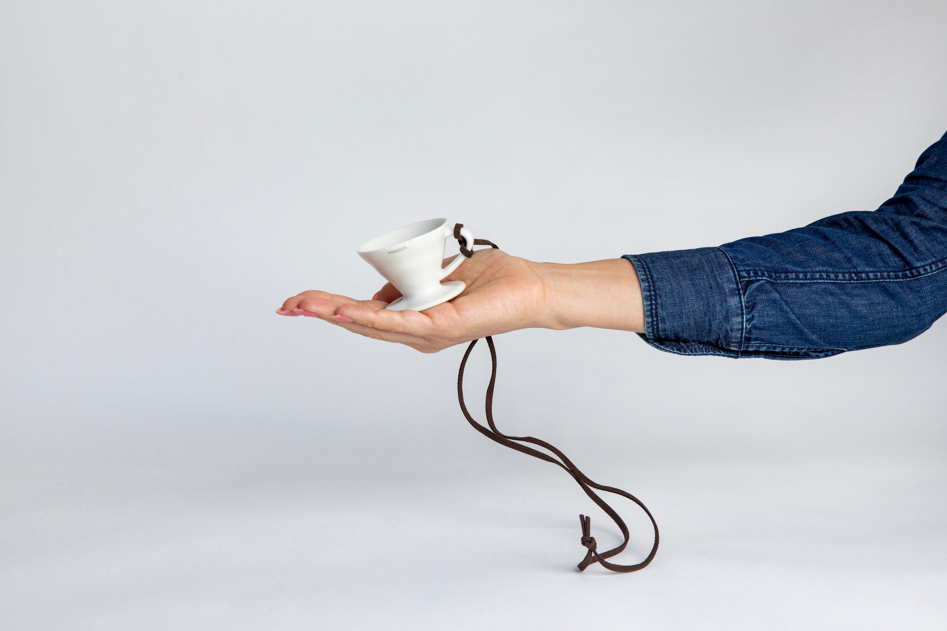 hand holding the white black cone shaped ceramic coffee dripper cup with handle and round base. with a brown rope tied on the handle, showcasing the size of the small cup can fit in the palm of a  hand. Set on white background