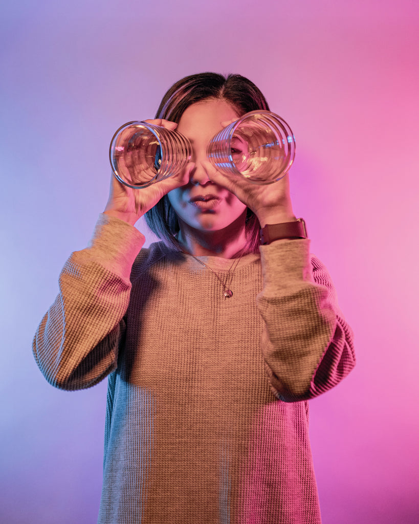 Model looking through two beehive shaped round double-wall coffee glasses, like binoculars or eye glasses, on a blue to pink gradient background.