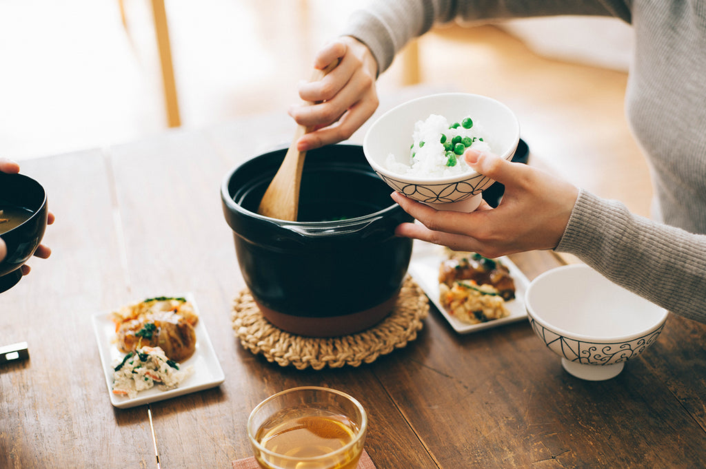 Woman serving freshly steamed white rice into a white bowl, holding white rice with green peas, from a black ceramic rice cooker with clay-colored powder coated bottom, glass lid, and black plastic lid knob.