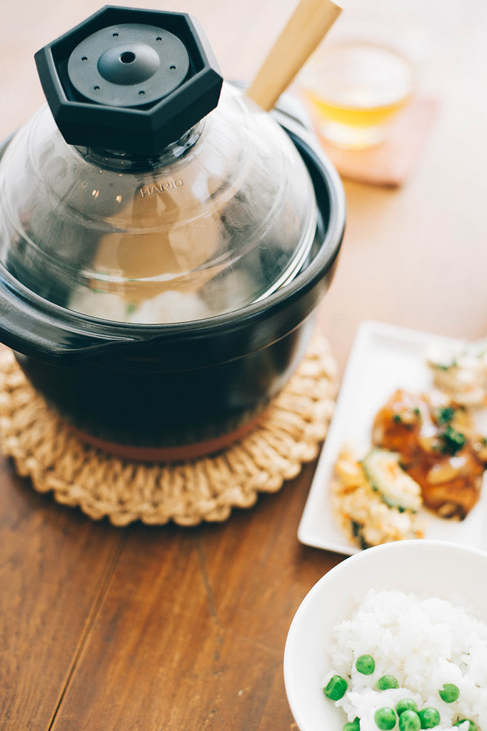 Black ceramic rice cooker with clay-colored powder coated bottom, glass lid, and black plastic lid knob. Rice cooker is holding a wooden rice serviing spoon and freshly steamed rice as it sits on a wood table with food.