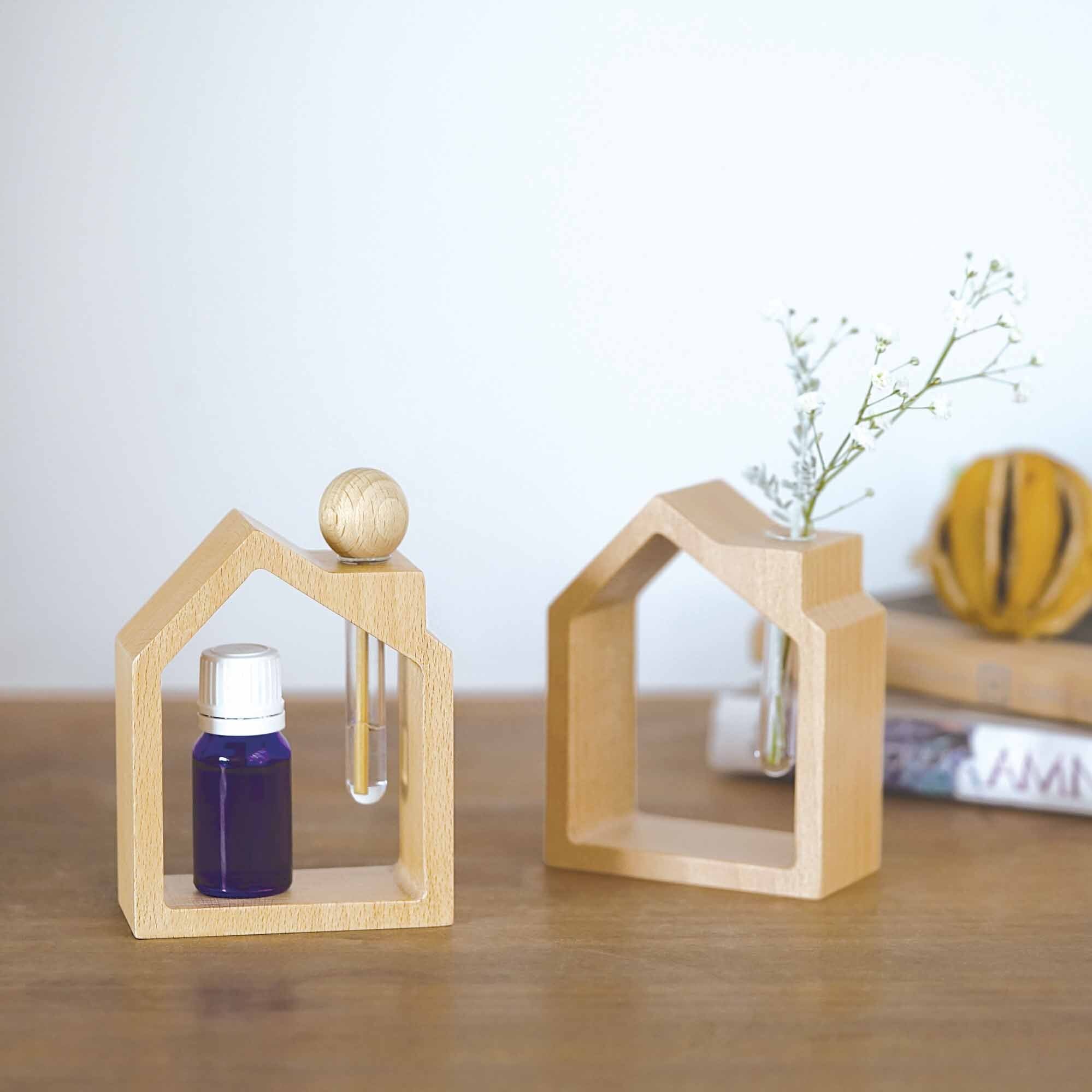 Two wooden house frames with wooden sticks shaped like a popsicle in front with small brown essential oils bottle and  pampas on the other house vile. Both houses set on a wood table with  two books and a light brown background.