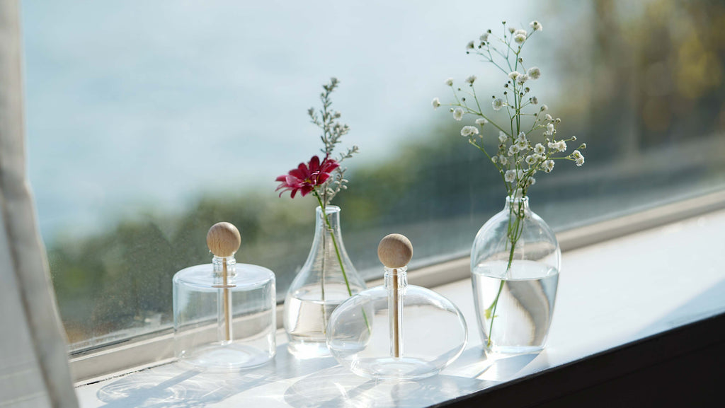 four glass diffusers of different shapes, cylindrical, tear-dropped, oval, and tuxedo, set on a white windowsill with blurred greenery outside the window. The cylindrical and oval shaped diffusers have the natural wood inserts in them and the remaining two diffusers have water and angel's breath flowers. A ruby red flower is also arranged in the tear drop shaped diffuser.