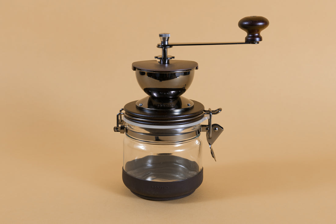 Round brown metal Coffee mill with handle, attached to a glass canister via hinge and latch with a rubber base on an orange backdrop.