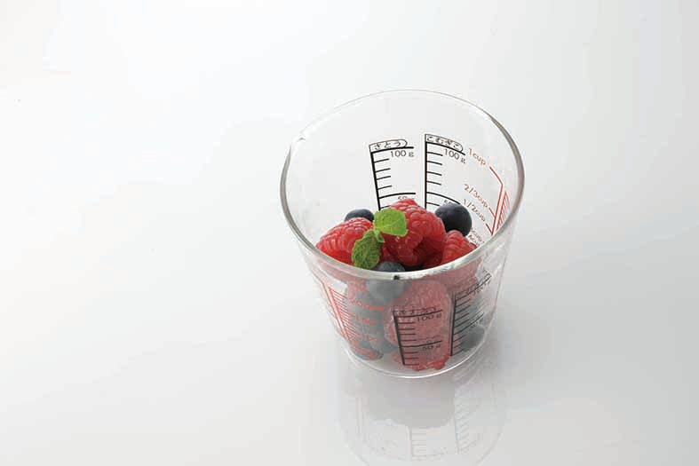 Glass measuring cup on white, reflective background with measurements in red and black on the inside and outside of the glass and partly filled with rasberries and blueberries.