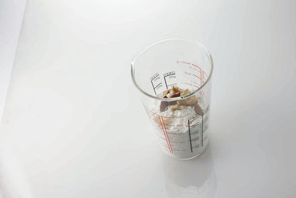 Tall glass measuring cup with small spout and red and black measurements on the inside and outside of the cup with flour and nuts inside on a white reflective background.