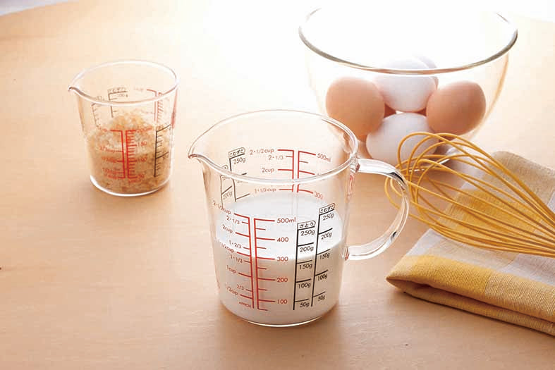 Glass measuring cup with spout, full glass handle, measurements in red and black on the inside and out, and filled with milk on a table setting with a small glass measuring cup with measurments in red and black on the inside and outside of the glass with bread crumbs inside. A yellow wisk on a folded yellow, white and beige checkered hand towel and a glass mixing bowl filled with brown and white eggs.