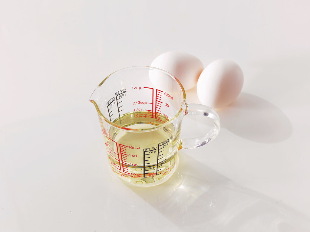 Glass measuring cup with spout, full glass handle, measurements in red and black on the inside and outside of the glass, and filled with a pale yellow oil and next to two white eggs on a white background.