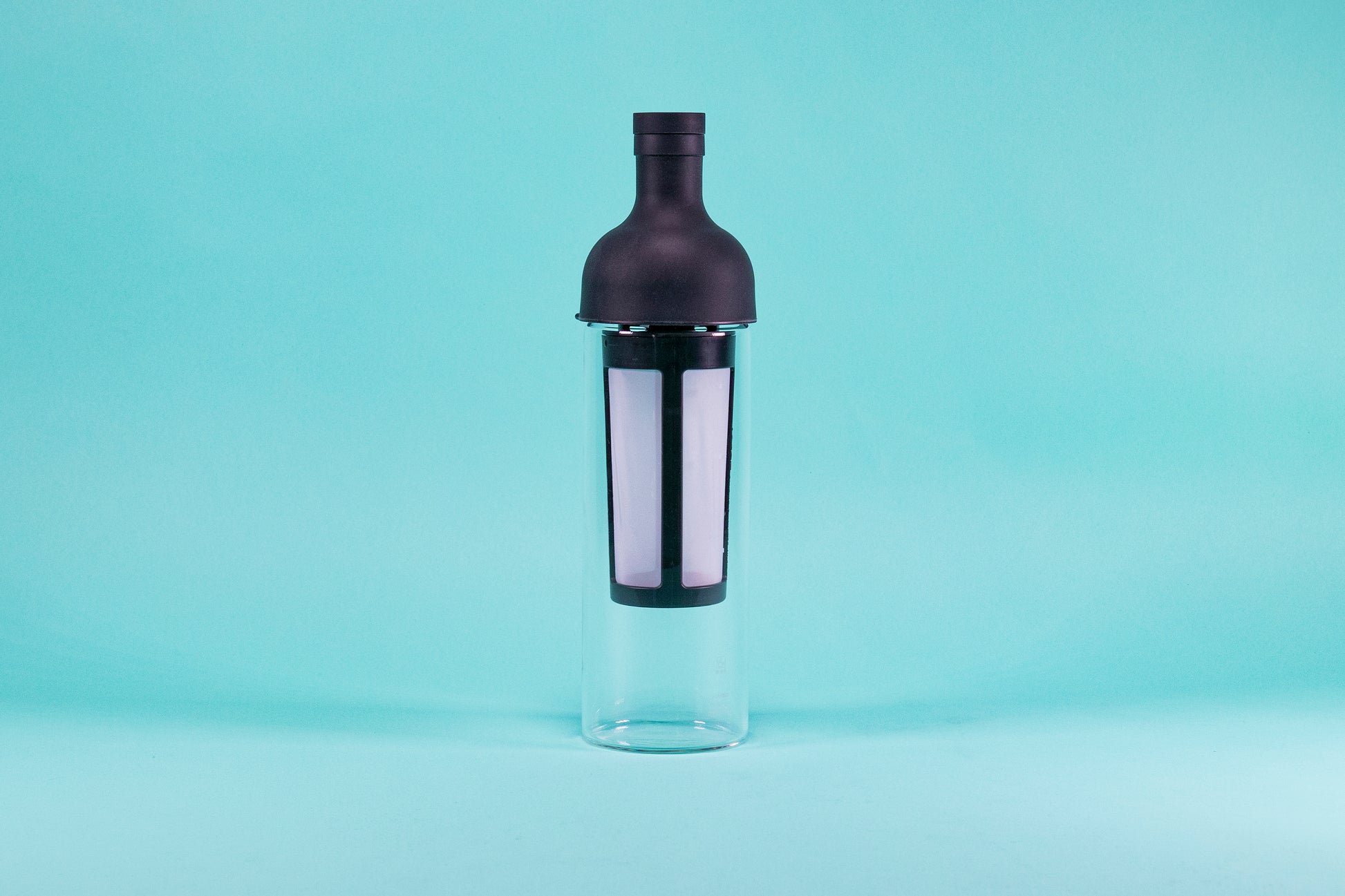 Tall glass container and white nylon mesh coffee filter, with black rubber wine bottle shaped top on a blue background.
